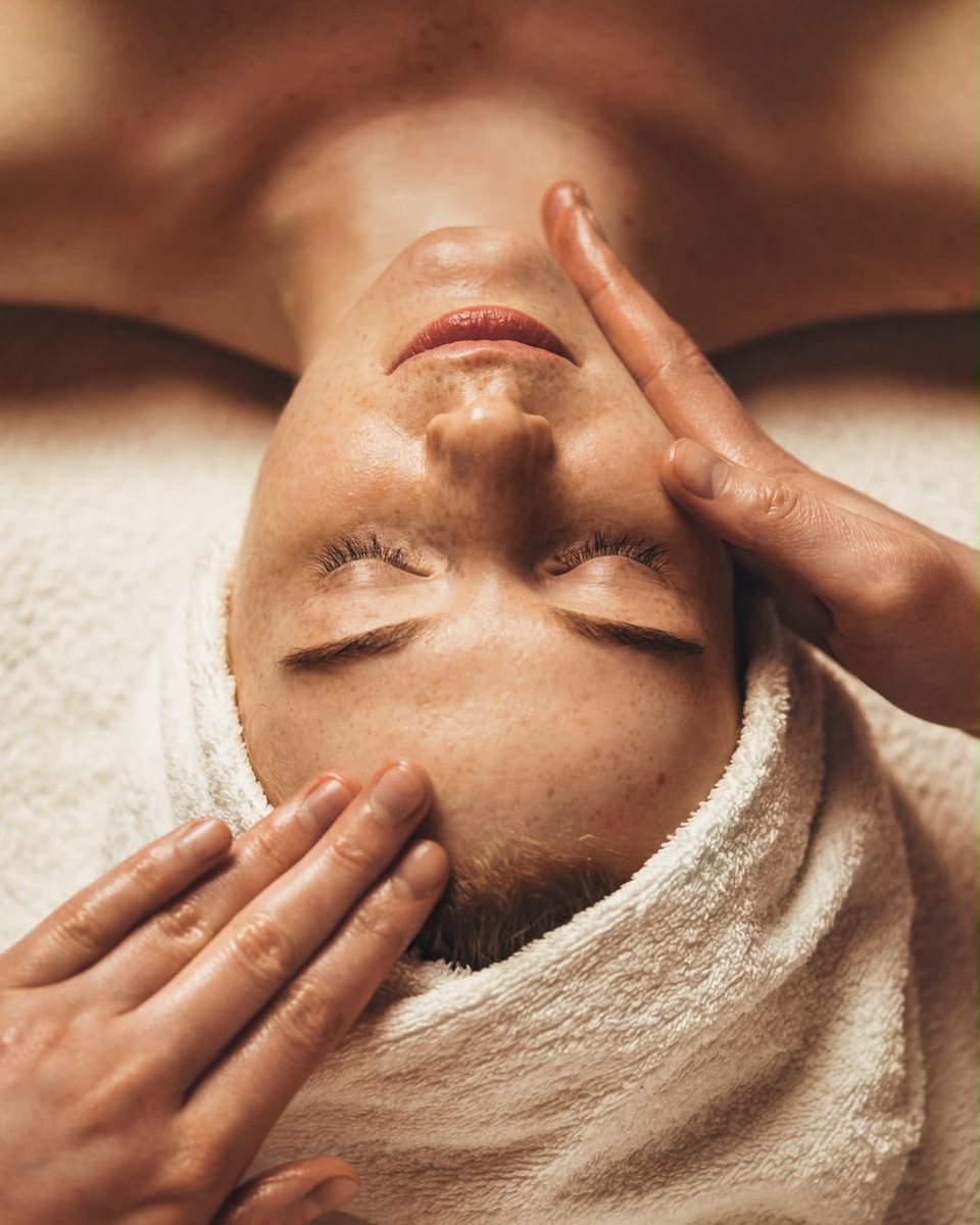 Warm, golden-lit facial massage with the client wrapped in a towel